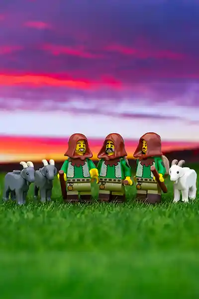 Three LEGO shepherd minifigures stand in a grassy field at sunset, each flanked by goats, beneath a vivid purple and orange sky.