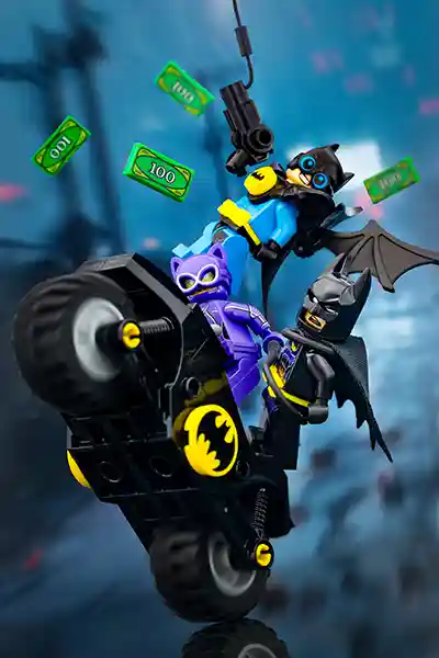 LEGO Batman, Robin (Nightwing), and Catwoman from The LEGO Batman Movie ride a Batcycle in high-speed action as $100 bills fly around them. Robin swings overhead with a grappling hook while Catwoman clings to the side, channeling the film's chaotic and comedic energy.