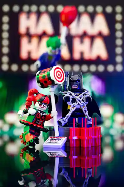 LEGO Harley Quinn prepares to smash a target with her mallet while Batman is chained and restrained on stage, looking distressed. In the background, the Joker gleefully waves a red balloon under glowing “HA HA HA” lights. Inspired by The LEGO Batman Movie, this scene captures the villains' theatrical flair and Batman's unlucky spotlight moment.