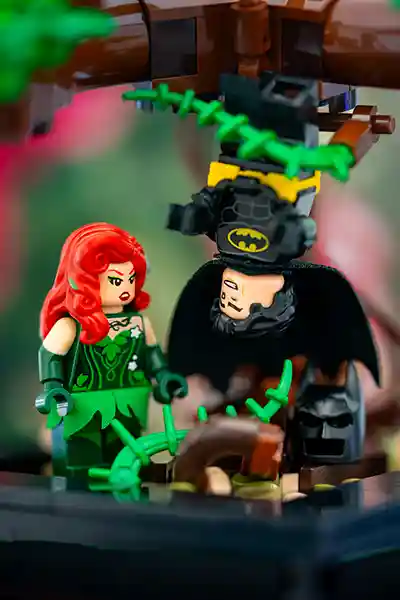 LEGO Poison Ivy glares at a captured Batman who is hanging upside down, ensnared in green vines from a tree. Batman's cowl has fallen off, revealing his unamused expression. The scene is a playful nod to The LEGO Batman Movie, highlighting Ivy's botanical powers and Batman's frequent predicaments.