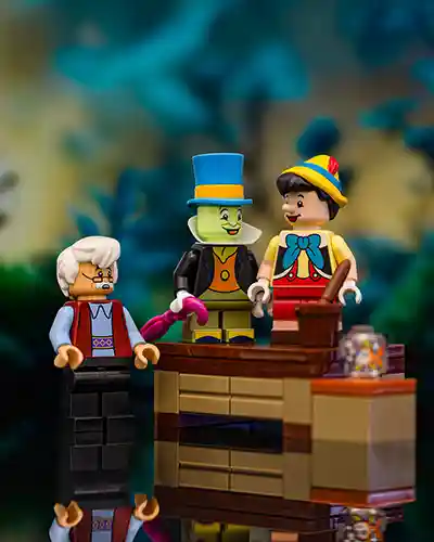 LEGO Pinocchio stands on a wooden workbench beside Jiminy Cricket and Geppetto. The scene captures the magical moment from Pinocchio where the puppet comes to life, with warm lighting and a cozy background suggesting Geppetto's workshop.