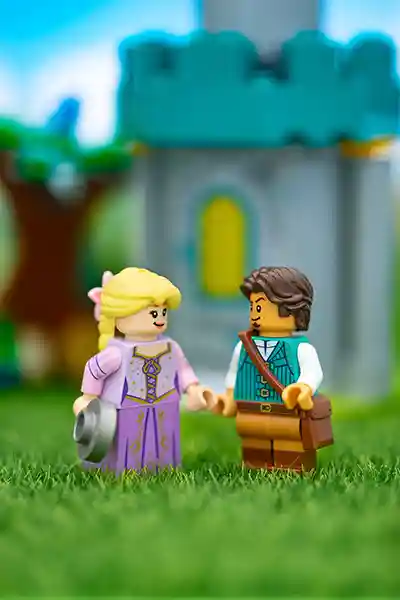 LEGO Rapunzel and Flynn Rider minifigures stand hand in hand on green grass in front of a castle tower, with trees and a bright blue sky in the background.