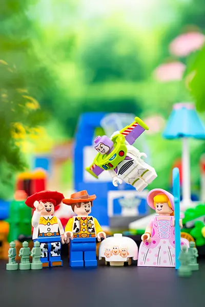 LEGO Toy Story minifigures of Woody, Jessie, Bo Peep, and Buzz Lightyear in a colorful outdoor setting, with Buzz mid-flight and green army men and sheep nearby.