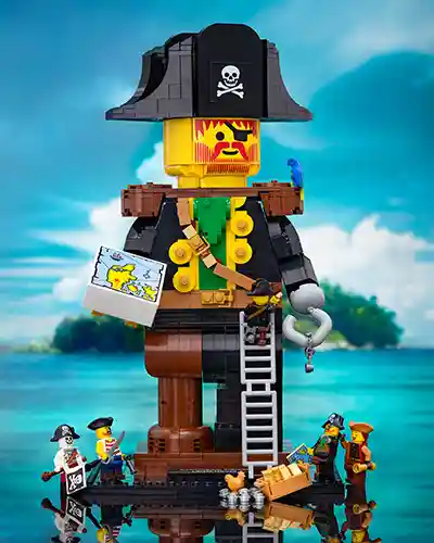 LEGO pirate minifigures build and explore around a giant pirate statue made from bricks, featuring an eye patch, hook hand, and a treasure map. The scene is set against a bright tropical ocean backdrop, with palm trees and an island in the distance.
