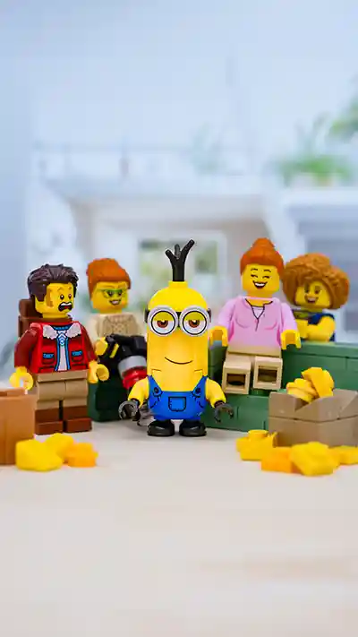 LEGO Minion figure stands proudly in the center of a bright living room setting, surrounded by laughing LEGO minifigures and scattered yellow bricks resembling bananas. The playful scene is a tribute to LEGO's April Fool's prank featuring a life-sized Minion build.