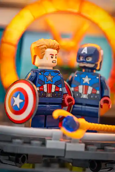 Two LEGO Captain America minifigures face each other in a tense standoff, one unmasked and one in full uniform, with Loki's glowing scepter lying in the foreground and a golden TVA time door glowing behind them.