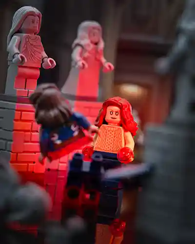 LEGO minifigures of Wanda Maximoff (Scarlet Witch) and Captain Carter face off in a dramatic scene surrounded by stone statues. Scarlet red lighting highlights Wanda's intense expression as she prepares a magical attack, with America mid-air, suspended by glowing energy.