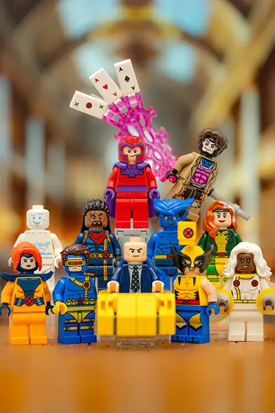 A group of LEGO X-Men minifigures is assembled in a dynamic pose on a smooth, reflective surface with a blurred grand hallway in the background. In the center, Professor X sits in his iconic yellow hoverchair, surrounded by team members including Cyclops, Wolverine, Storm, Jean Grey, Beast, Rogue, Iceman, and Bishop. Magneto hovers behind them with a determined expression, while Gambit dramatically throws energized playing cards, represented by translucent pink energy effects. Each minifigure is detailed with their signature costumes and expressions, capturing the essence of the beloved superhero team.
