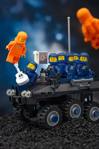 A team of dark blue LEGO Classic Space astronauts rides a black moon rover while one plays a white guitar. Two orange astronauts float nearby in zero gravity. A Classic Space flag waves from the vehicle under a starry space sky.