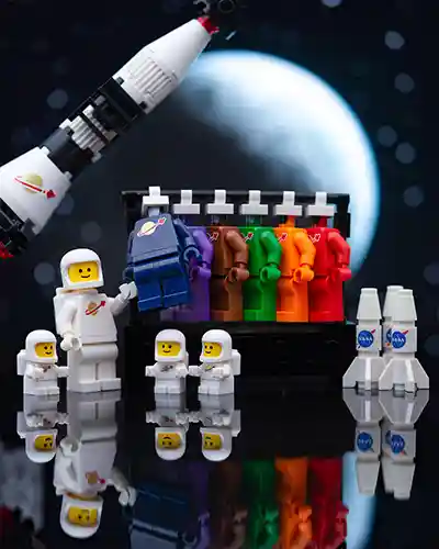A white LEGO Classic Space minifigure unveils a new dark blue spacesuit in front of a rainbow lineup of other suits. Three micro astronauts stand proudly nearby with two NASA-style rockets and a moonlit backdrop.