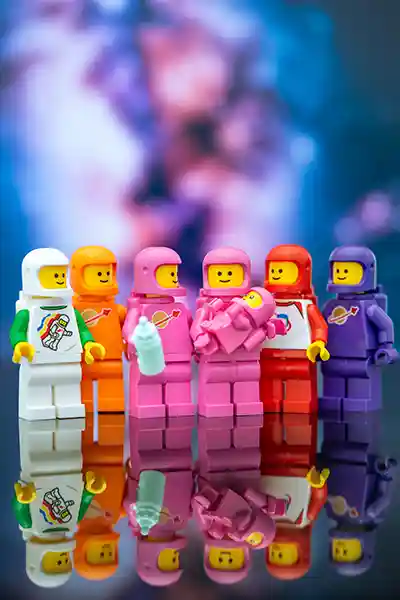 Colorful LEGO astronauts gather around two pink-suited figures holding a baby and bottle, celebrating under a cosmic, dreamy backdrop.
