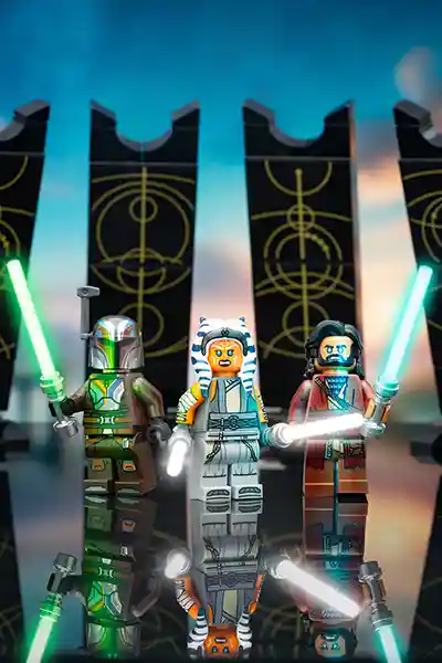 Three LEGO Star Wars minifigures—Sabine Wren, Ahsoka Tano, and Baylan Skoll—stand ready with lit lightsabers in front of tall black stone pillars etched with golden circular patterns, under a vivid twilight sky.