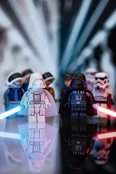 Two LEGO Darth Vader minifigures stand side by side—one in white with a blue lightsaber, the other in black with a red lightsaber. Rebel and Imperial troops are blurred in the background, with dramatic light rays splitting the scene down the middle.