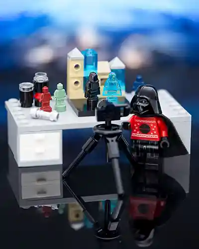 LEGO Darth Vader wears a festive red sweater while standing behind a camera setup on a tripod. He's filming a miniature diorama featuring tiny Star Wars holograms and minifigures on a desk, surrounded by props like lights, mugs, and canisters. The scene humorously blends Sith seriousness with seasonal cheer.