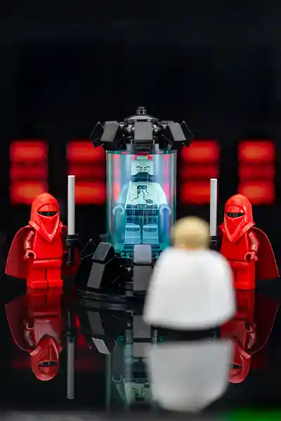 LEGO Darth Vader rests inside a Bacta tank, flanked by two red Imperial Guards, as a white-caped figure approaches in a dark Imperial chamber with glowing red control panels.