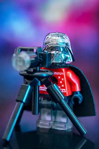 Close-up of LEGO Darth Vader wearing a festive red holiday sweater, standing behind a black camera on a tripod. The background is a vibrant blend of purple, blue, and pink hues, giving a colorful contrast to the Sith Lord’s serious expression and cheerful attire.