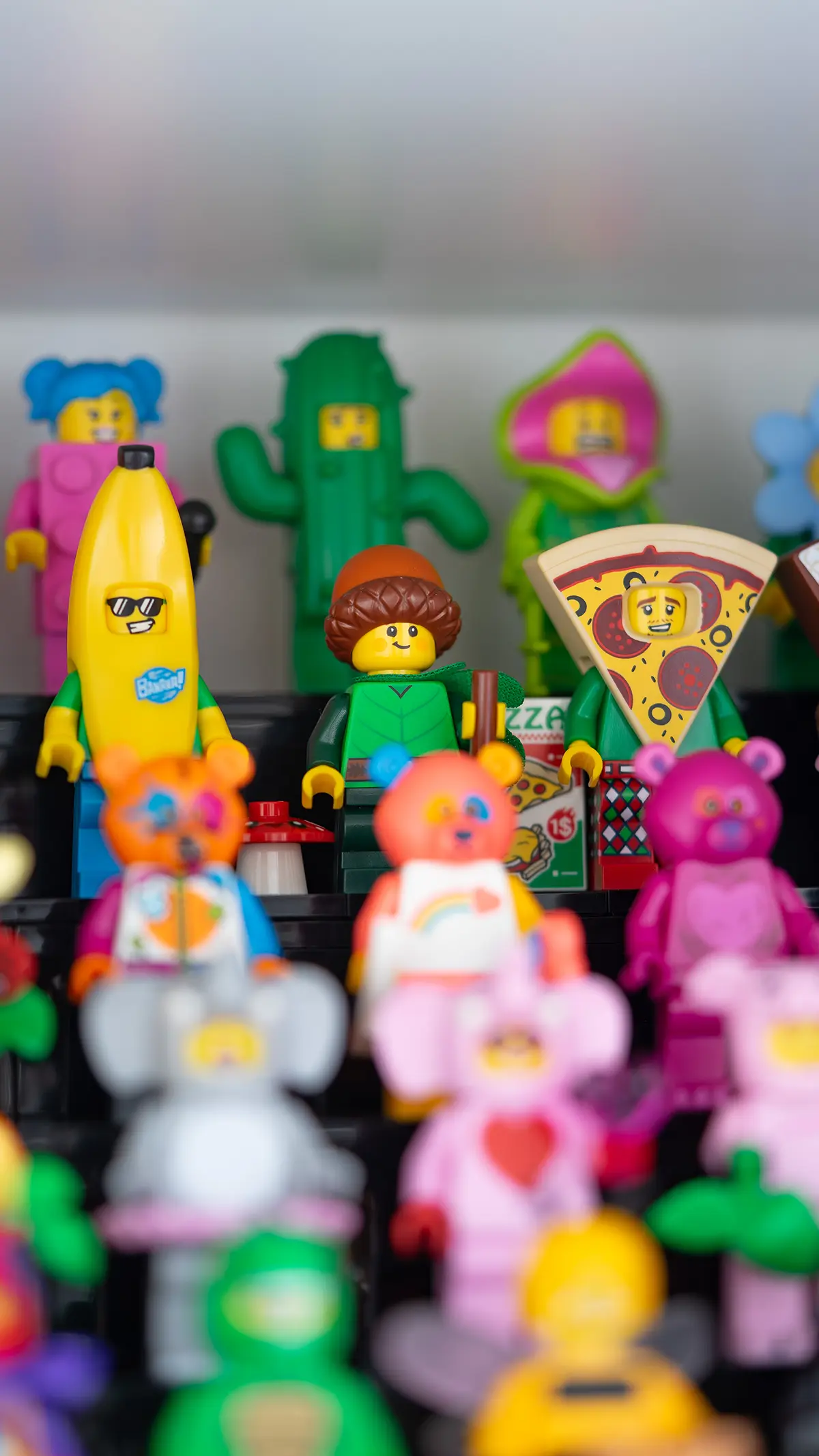 LEGO minifigures in whimsical outfits including a banana suit, a tree costume, and a pizza slice, with colorful costume characters and flowers in the background.