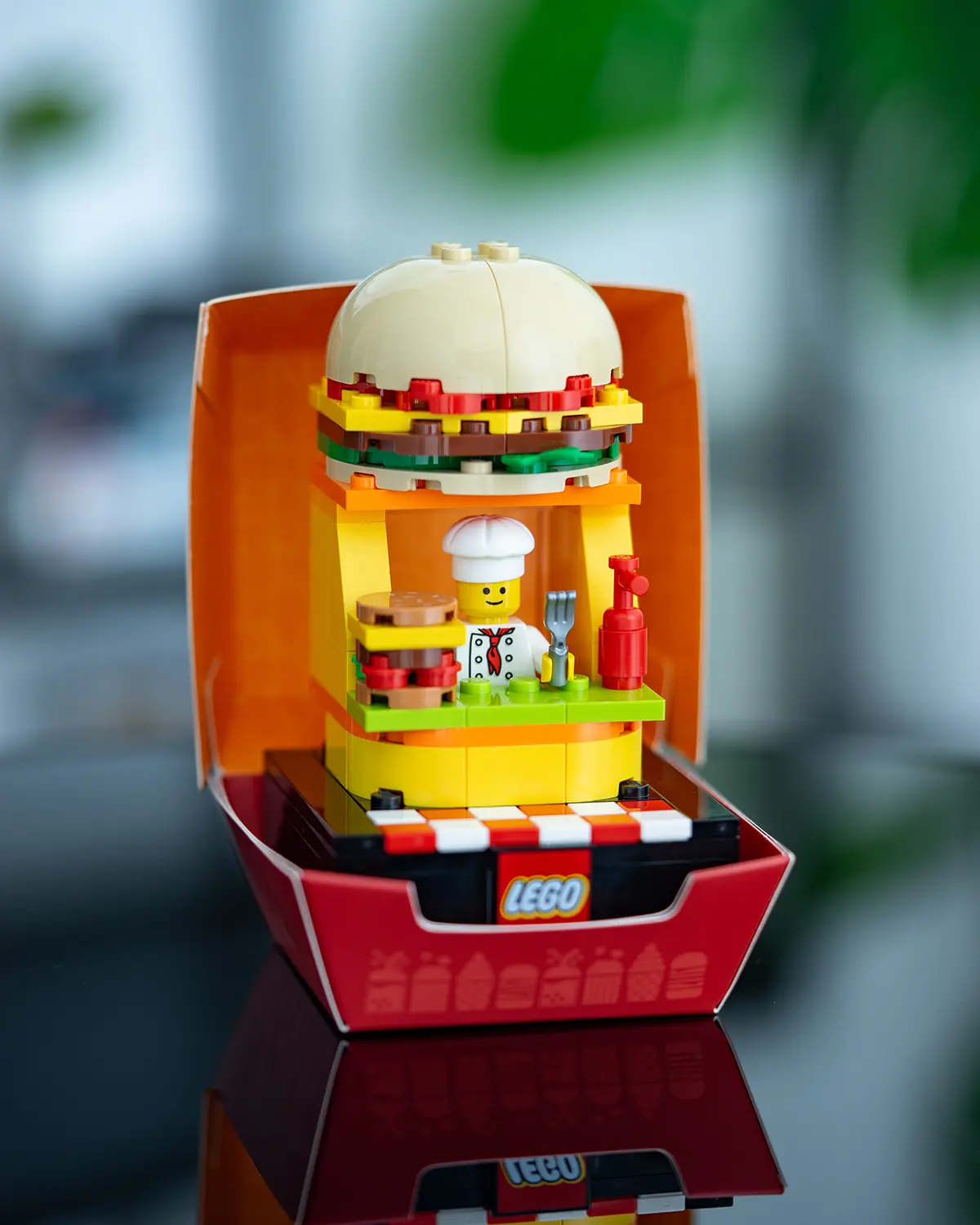 LEGO burger-themed kiosk with a large brick-built hamburger bun on top, a smiling chef minifigure, and condiments on the counter.