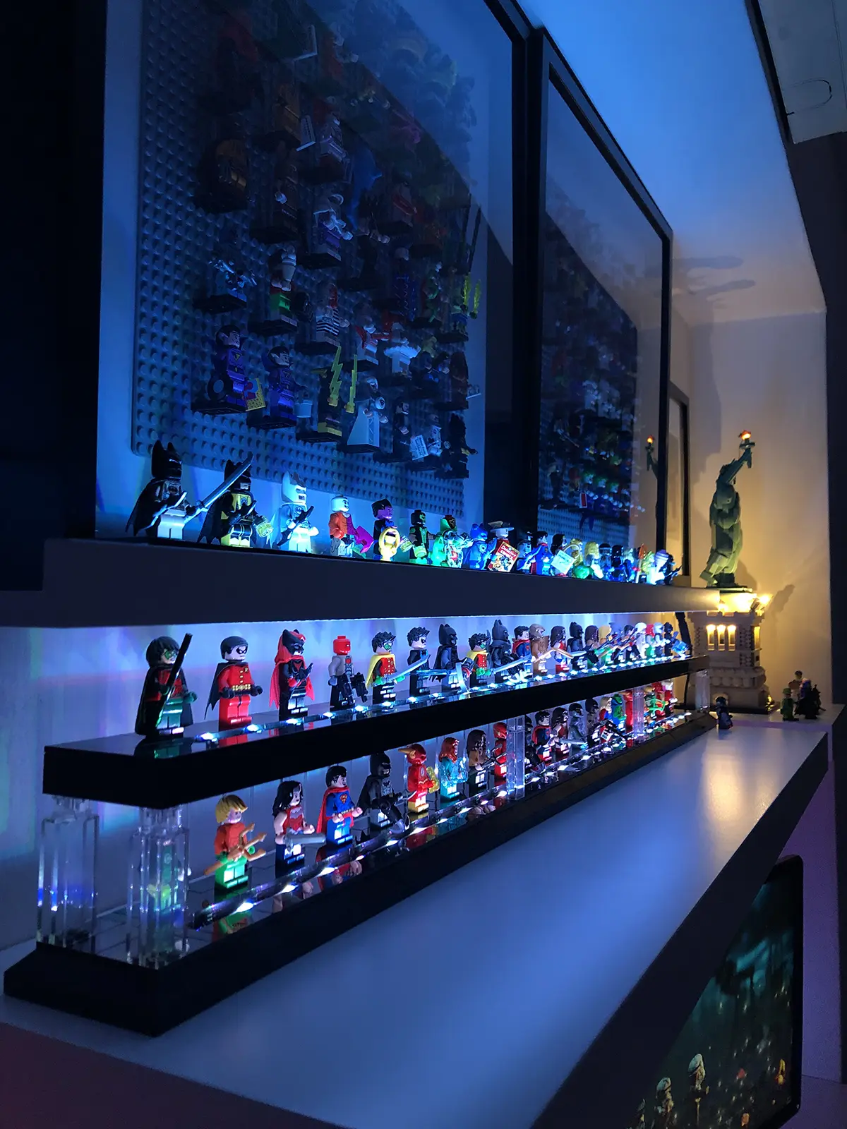 LEGO superhero minifigures displayed on LED-lit shelves, glowing in blue and white light. The setup features DC characters like Batman, Superman, and Wonder Woman, with framed minifigure displays and a LEGO Statue of Liberty build in the background.