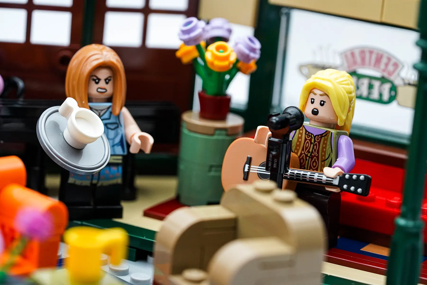 LEGO minifigures of Rachel and Phoebe in Central Perk; Rachel is angrily holding a tray while Phoebe sings into a mic with her guitar