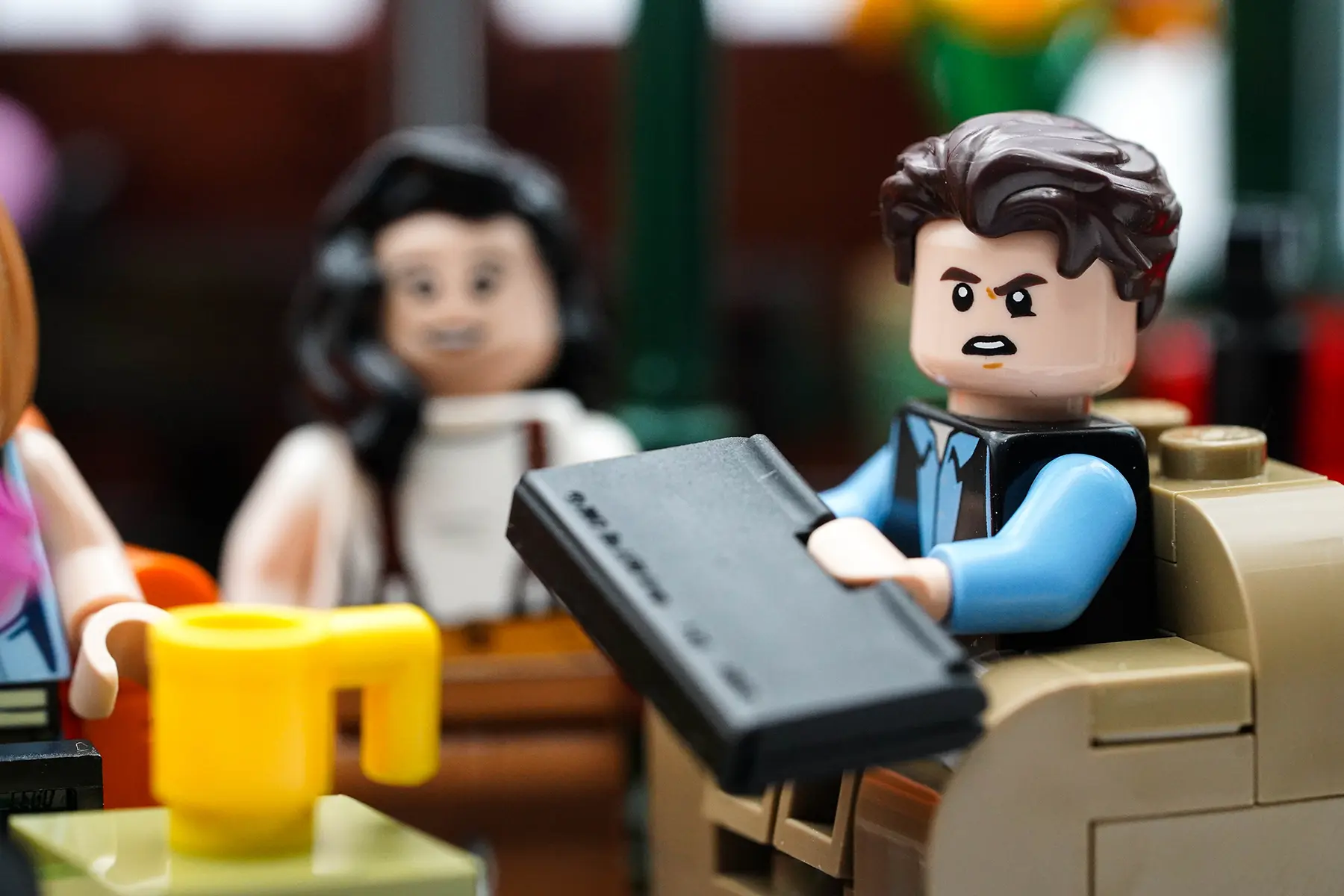 LEGO Ross Geller holding a laptop with an upset expression while Monica smiles in the background at Central Perk.
