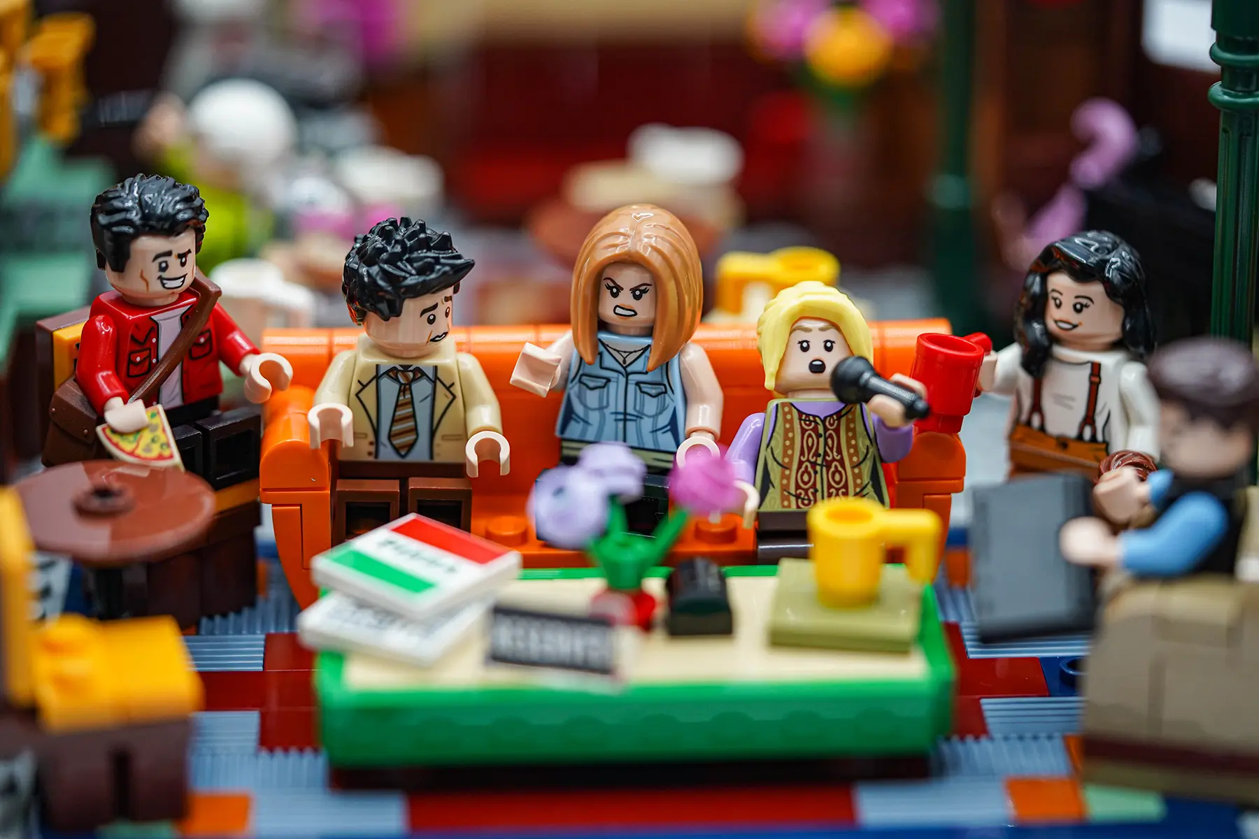 A LEGO Central Perk coffee table with flowers, pizza boxes, and a “Reserved” sign, with the Friends blurred in the background.