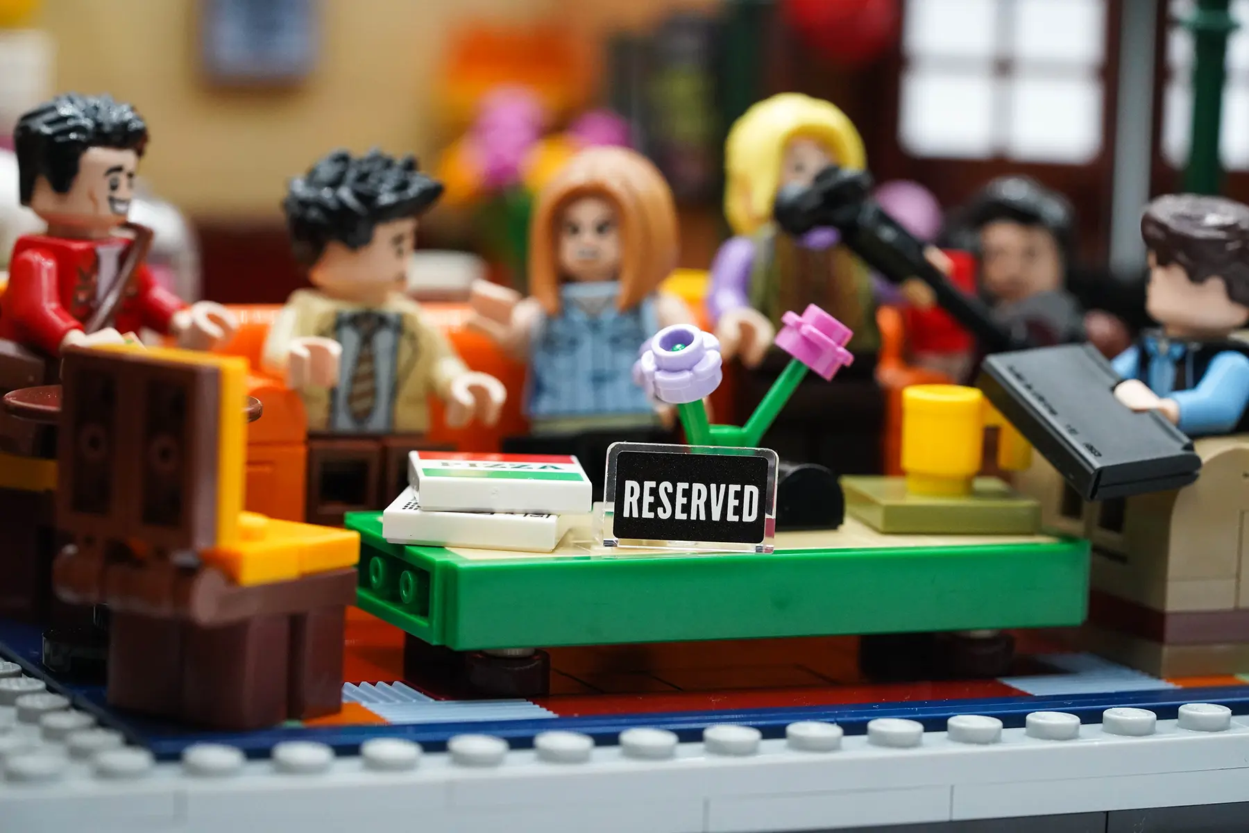 A LEGO Central Perk coffee table with flowers, pizza boxes, and a “Reserved” sign, with the Friends blurred in the background.