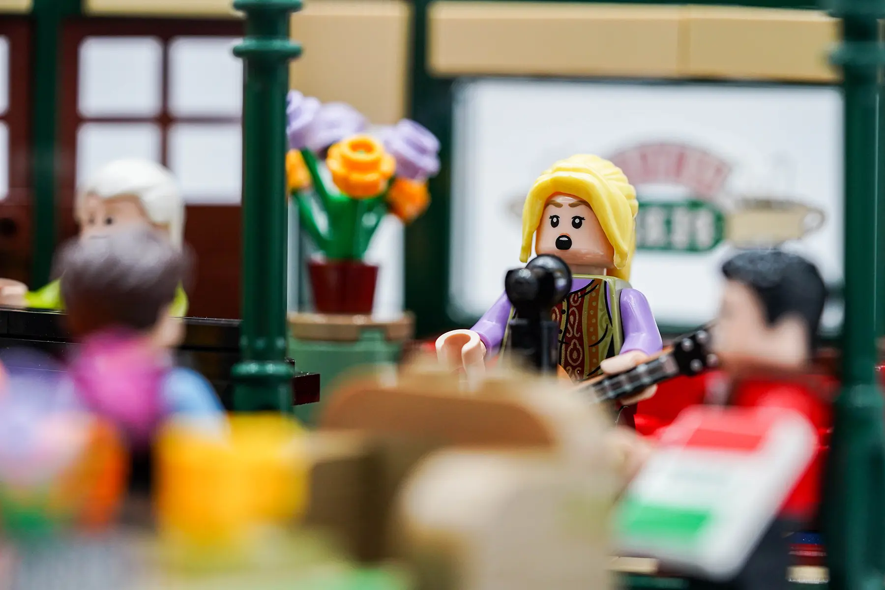 Phoebe singing into a mic with her guitar at LEGO Central Perk as other minifigures watch from the background.