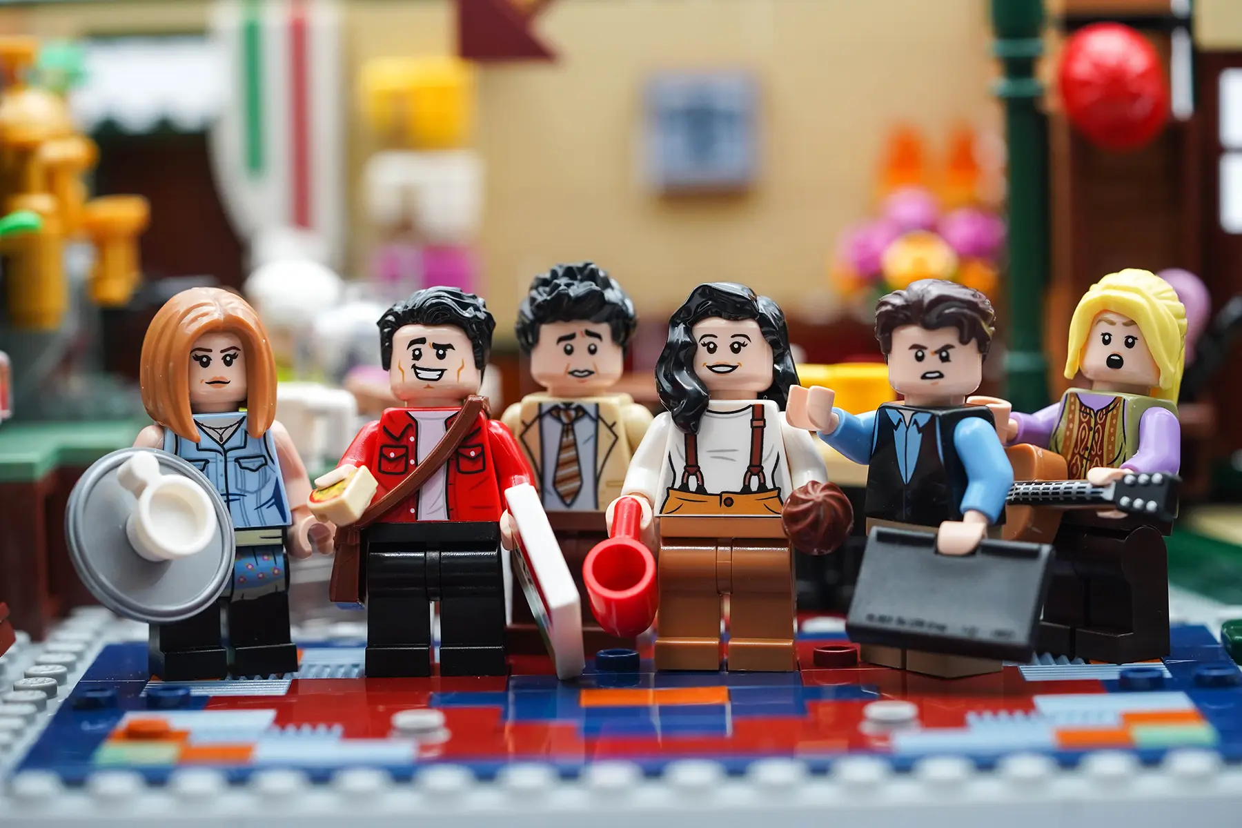LEGO minifigures of all six Friends characters posing together in front of the Central Perk backdrop, each holding signature items.