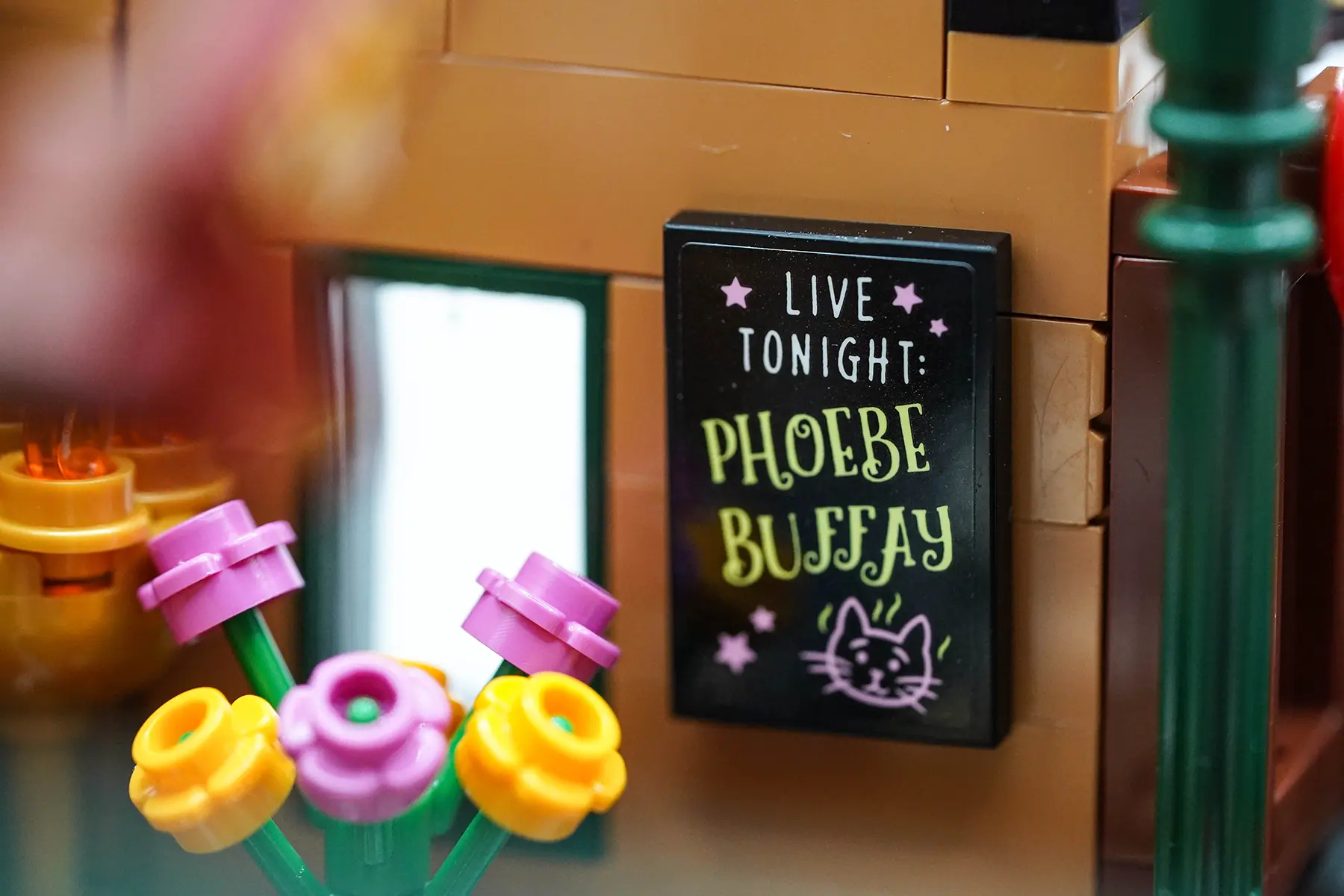 Close-up of a LEGO poster in Central Perk that reads “Live Tonight: Phoebe Buffay” with a drawing of a cat, surrounded by flowers and warm lighting.