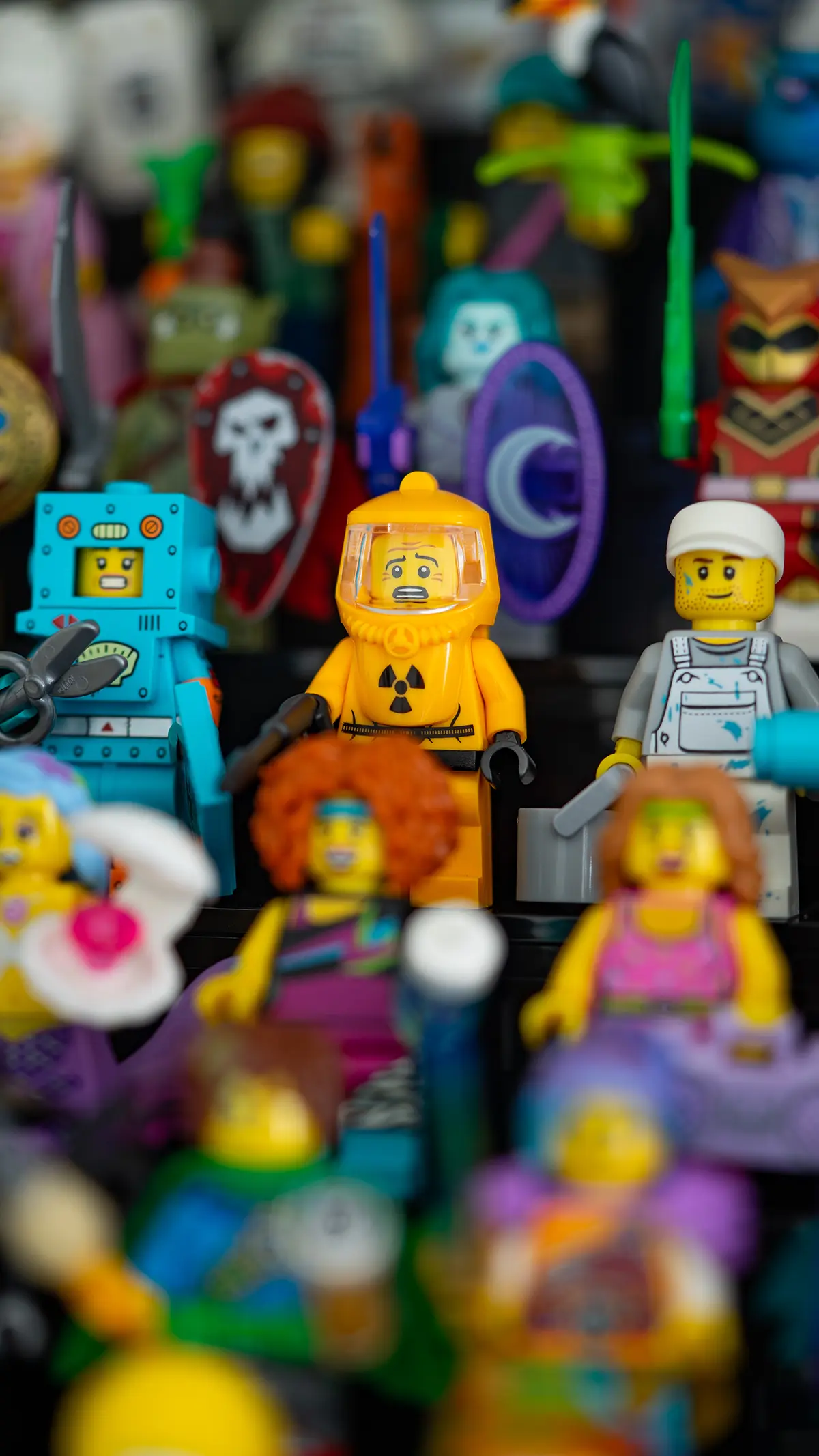A LEGO minifigure collection featuring a hazmat suit character, a painter, a robot, and other sci-fi and fantasy-themed figures in the background.