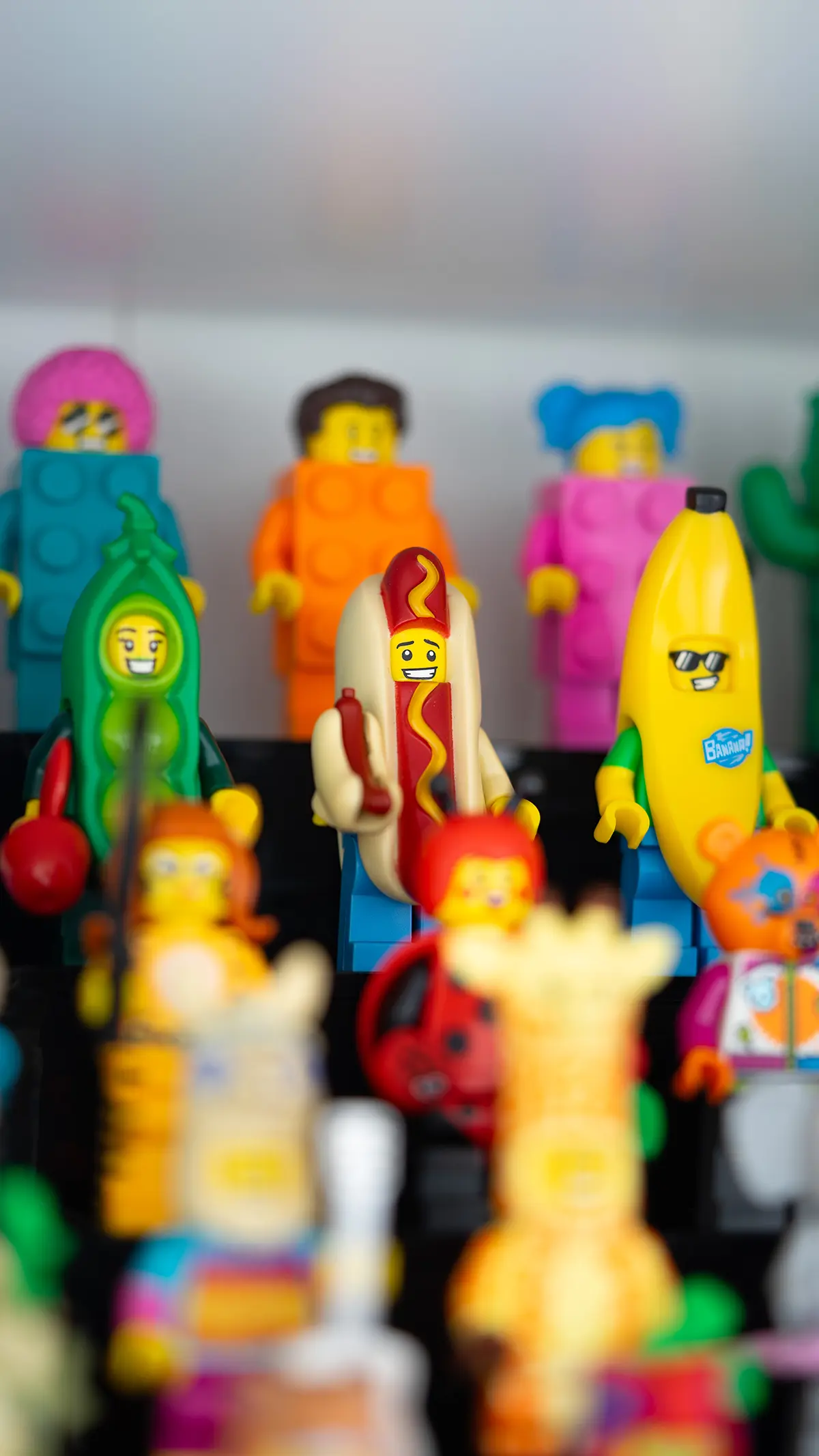 A display of LEGO minifigures dressed in food and costume-themed outfits, including a hot dog, banana, and pea pod, with brightly colored brick-suit minifigures in the background.