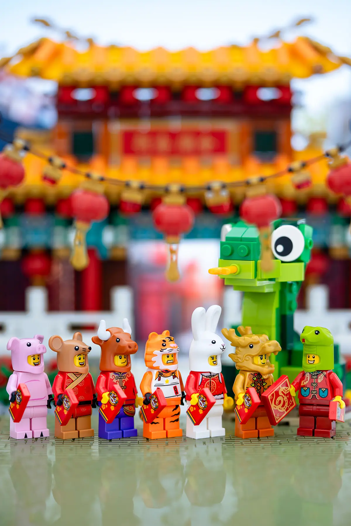 A festive lineup of LEGO minifigures dressed as Chinese zodiac animals, including the pig, rat, ox, tiger, rabbit, dragon, and the newly introduced snake. Each character holds a red envelope while standing in front of a traditional Chinese temple decorated with red lanterns. A playful brick-built green snake with wide eyes stands prominently on the right, symbolizing the Year of the Snake.