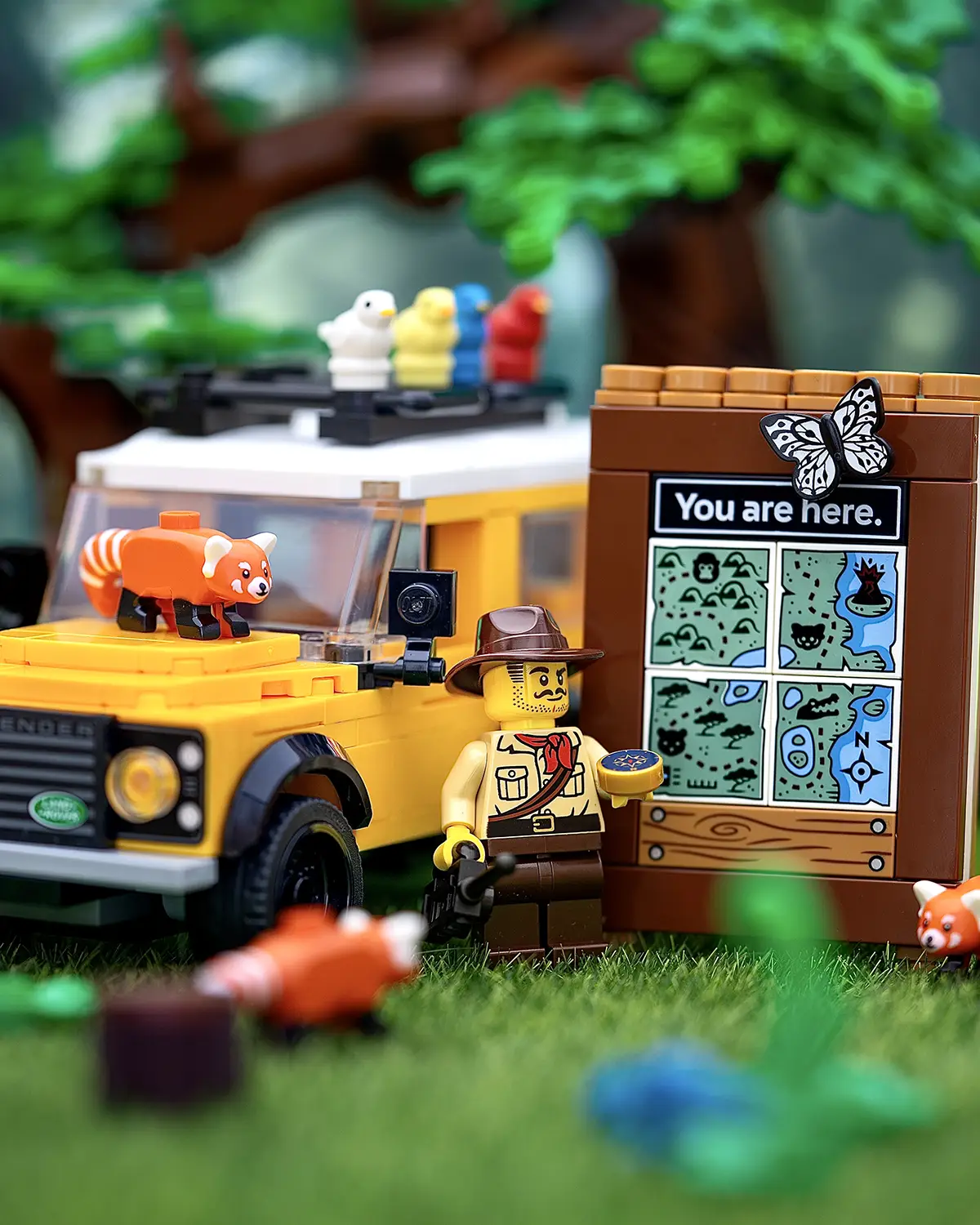 A LEGO park ranger with a compass stands next to a nature signboard featuring four connected printed tiles that form a seamless illustrated map, with a yellow Land Rover, red pandas, colorful birds, and forest scenery in the background.