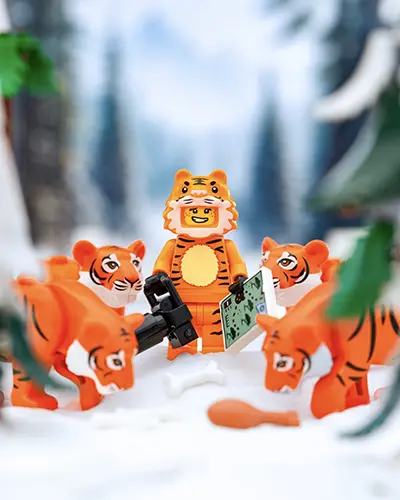 LEGO minifigure in a tiger costume stands in snow surrounded by five LEGO tiger animals, holding a camera and a map, with forest scenery in the background.