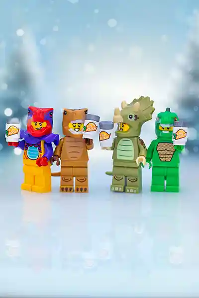Four LEGO minifigures in colorful dinosaur and animal suits—red lizard, brown T-Rex, green triceratops, and green crocodile—stand side by side smiling and holding Vita Rush drinks, set against a soft wintery bokeh background.