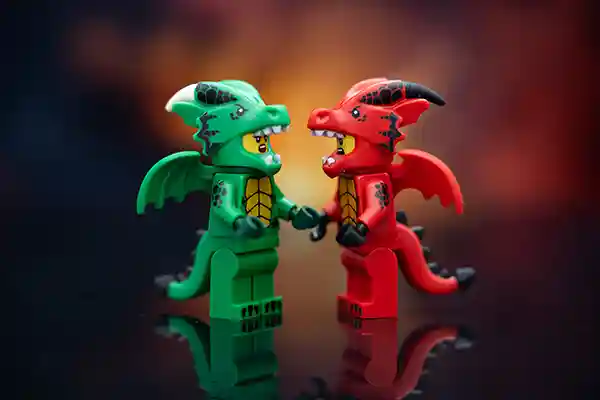 Two LEGO minifigures in detailed dragon suits—one red, one green—stand face to face on a reflective surface with a fiery gradient background, seemingly locked in an intense but playful stare.