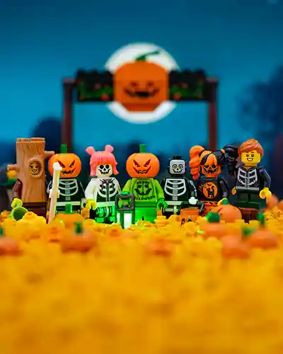 A festive group of LEGO minifigures dressed in skeleton and pumpkin-themed costumes gather in a glowing orange pumpkin patch under a full moon with a smiling jack-o'-lantern sign in the background.