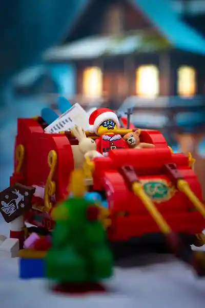 A LEGO Santa minifigure snoozes in a red sleigh packed with gifts, cuddled up with a teddy bear and bunny, while a snowy cabin glows warmly in the background.