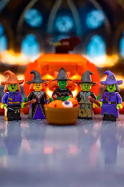 Five LEGO witches with green skin and colorful outfits stand in front of a glowing jack-o'-lantern, with a basket of spooky treats including a giant eyeball.