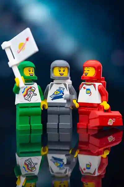 Three LEGO minifigures in green, gray, and red classic space suits share a joyful moment, holding a flag, wrench, and toolbox against a cosmic backdrop.